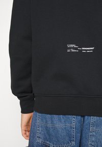 Sweat-shirt noir avec ourlet et poignets côtelés, arborant un texte blanc détaillé au dos. Le tissu semble doux avec une texture lisse.