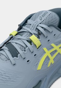 Lys grå idrettssko med mesh-overdel, gule detaljer og teksturert design. Har snører og en støttende hældesign.