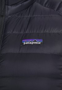 Svart puffjacka med kviltat mönster, med en Patagonia-logotyp med bergsgrafik och färgglada ränder. Slät textur.