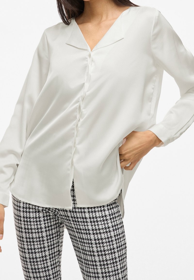 Blouse blanche en tissu doux et soyeux avec un col en V, des manches longues et une patte de boutonnage sur le devant. Portée avec un pantalon à motif pied-de-poule noir et blanc.