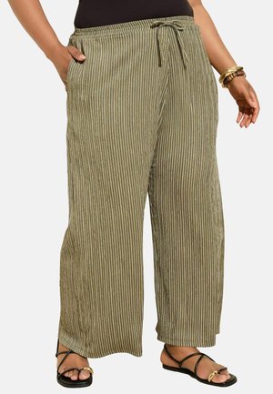 Femme portant un pantalon large vert olive avec des rayures blanches verticales, des sandales noires, un haut noir et des bracelets à un poignet.