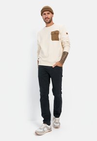 Beige trui met een bruine borstzak, gecombineerd met donkere broeken en grijze sneakers. De trui heeft een ronde hals en ribbelmanchetten.