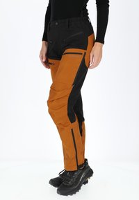 Pantaloni outdoor impermeabili con una fascia nera in vita, gambe arancioni con dettagli neri, numerose tasche con zip e vestibilità affusolata.