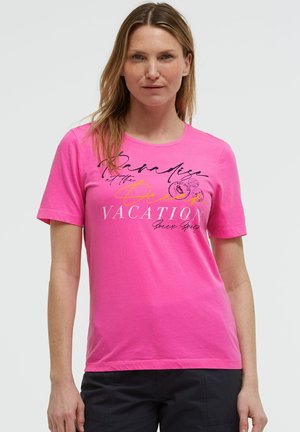 Frau trägt ein hellpinkes T-Shirt mit kurzen Ärmeln und schwarzem Text sowie Grafiken, kombiniert mit dunklen Hosen, steht vor einem schlichten Hintergrund.