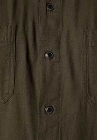 Camicia verde oliva con bottoni a pressione, caratterizzata da un tessuto texturizzato, bottoni neri e due tasche sul petto con alette appuntite.