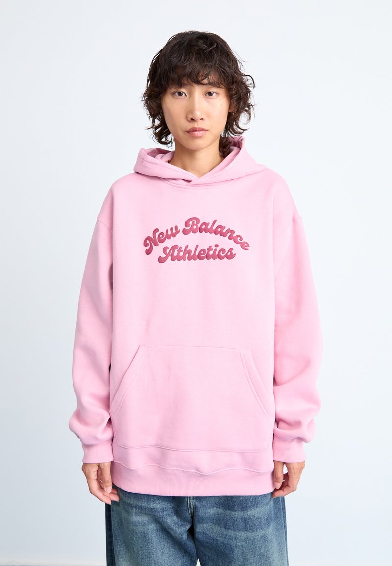 Jeune personne avec des cheveux bouclés portant un sweat à capuche New Balance Athletics rose clair et un jean bleu ample, sur un fond simple et clair.