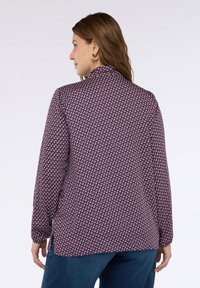 Femme aux cheveux bruns portant des boucles d'oreilles en or et un chemisier à motifs à manches longues associé à un pantalon bleu foncé, de dos.