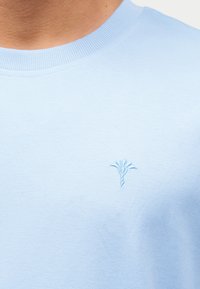 Ανοιχτό μπλε μπλουζάκι με στρογγυλή λαιμόκοψη και μικρό κεντημένο λουλουδάτο λογότυπο στο αριστερό μέρος του στήθους, που φοριέται από άτομο.