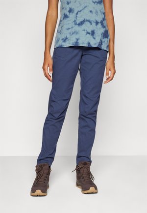 Marineblaue Hosen aus leichtem Material, mit schmaler Passform und elastischen Bündchen, kombiniert mit braunen Schnür-Wanderschuhen.