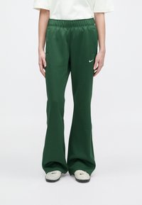 Cilvēks, ģērbies tumši zaļos flared sporta biksēs ar sānu kabatām un baltu logo tuvu kabatai, apģērbts ar baltām kedām bez auklām ar melniem detaļām.