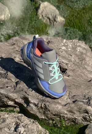 Chaussure de trail en gris, bleu et violet avec des lacets aqua reposant sur un terrain rocheux avec de l'herbe en arrière-plan.