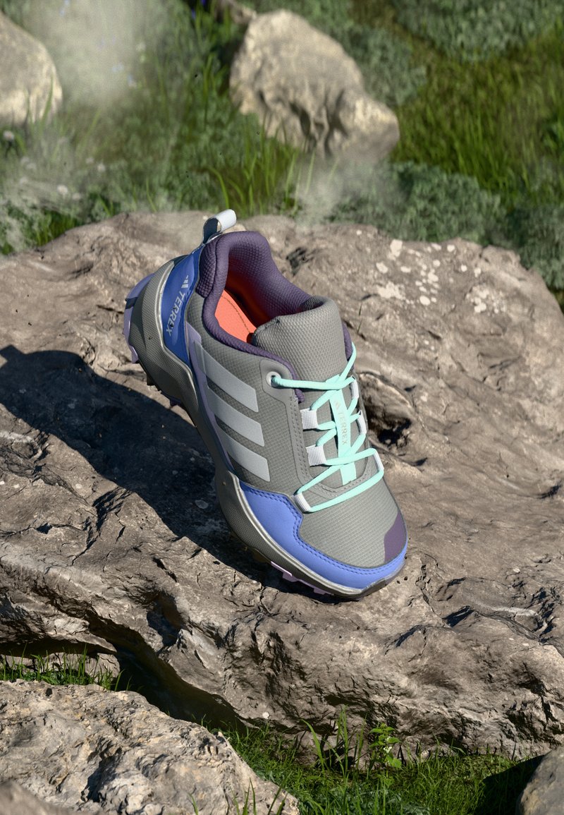 Sapatilha de trail running em cinza, azul e roxo com atacadores aqua repousando em terreno rochoso com relva ao fundo.