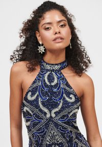 Námořnické modré šaty s halter krkem zdobené složitým stříbrným a modrým korálkováním, s přiléhavým střihem a flitrovou texturou.