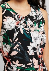 Černé šaty bez rukávů s květinovým vzorem, který zahrnuje bílé lilie, růžové akcenty a zelené listy; výstřih ve tvaru v.