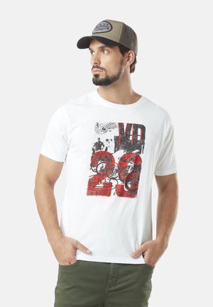 T-shirt blanche avec un imprimé graphique présentant des accents rouges et noirs. La personne qui la porte l'associe à une casquette grise, arborant un logo sur le devant.