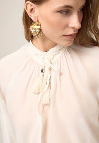 Blusa de suave tejido crema, con un cuello alto, lazos decorativos y borlas. Un pendiente de aro texturizado en oro añade un acento audaz.