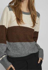 Sweter z dzianiny z poziomymi paskami w odcieniach beżu, brązu i szarości. Okrągły dekolt, ściągacze na rękawach i miękka tekstura. Noszony z czarnymi spodniami.