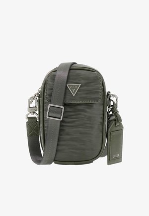 Sac bandoulière compact vert foncé texturé avec sangle ajustable, garniture argentée, plaque logo Guess et étiquette bagage attachée.