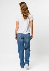 Žena s dlhými kučeravými vlasmi, oblečená v bielom tričku s krátkym rukávom, modrých džínsoch a bielych teniskách, stojaca pred bielym pozadím.