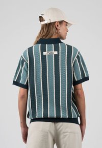 Mujer con un suéter de manga corta a rayas verticales azul y blanco con cuello y una gorra beige, de espaldas sobre un fondo gris liso.