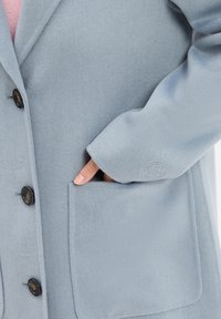 Cappotto in misto lana azzurro chiaro con bottoni neri, tasca laterale e logo impresso sulla manica. Tessuto morbido e design su misura.