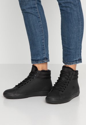 Personne portant des baskets montantes noires doublées de fourrure et un jean bleu, debout sur un sol gris clair, jambes croisées.