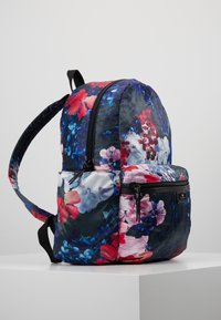 Reppu, jossa punaisen, sinisen, violetin ja valkoisen kukkakuvio, mustat vetoketjut ja pehmustetut olkahihnat, seisoo valkoisella pinnalla.