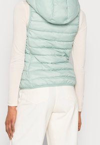 Light mint green padded vest with a hood, featuring horizontal quilted pattern and elastic hem, worn over a long-sleeve cream top and white pants.