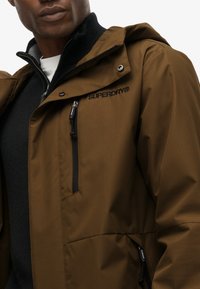 Brązowa wodoodporna kurtka z zamkiem z przodu, kapturem i wieloma zamykanymi na zamek kieszeniami. Posiada czarną logo na klatce piersiowej oraz teksturowany materiał.