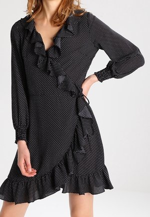 Kobieta w czarnej sukience z kopertowym wiązaniem w białe groszki, z długimi rękawami, falbanianym kołnierzem i dołem, pozująca z jedną ręką na biodrze.