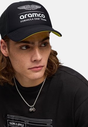 Giovane uomo con capelli di media lunghezza indossa un cappellino nero Aston Martin Aramco Formula One, una maglietta nera e una catena d'argento con due ciondoli.