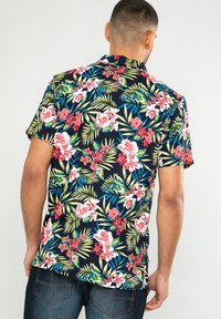Hombre vestido con una camisa de manga corta color marino con patrones florales tropicales coloridos y unos jeans azul oscuro, de espaldas contra un fondo sencillo.