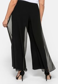 Černé volné kalhoty s průhledným overlayem, elastickým pasem a detailem rozparku vzadu. Spárované s vysokými podpatky. Hladká textilie.