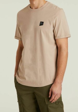 Mężczyzna ubrany w beżowy t-shirt z krótkim rękawem i okrągłym dekoltem z małym czarnym logo na piersi, w połączeniu z oliwkowo zielonymi spodniami typu cargo.