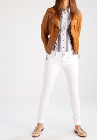 Brun lädermotorcykeljacka, grårandig skjorta med knappar, vita slim fit jeans och beige mockastilskor med fransdetaljer.