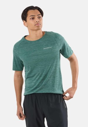 Mand i grøn kortærmet sportstrøje og sorte shorts, der justerer trøjens kant, stående mod en ensartet lys baggrund.