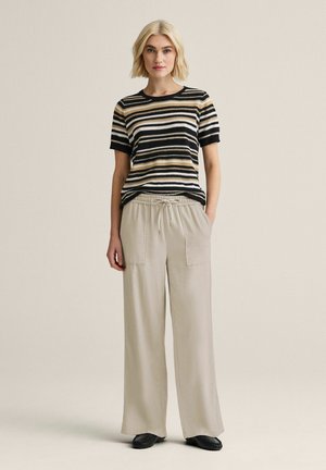 Kvinde stående iført en kortærmet sort, beige og hvidstribet sweater, beige bukser med vide ben og sorte sko, neutral baggrund.