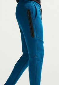 Μπλε παντελόνι jogging με εφαρμοστή σχεδίαση, κατασκευασμένο από μαλακό ύφασμα. Διαθέτει μαύρη τσέπη με φερμουάρ στο πλάι και διακριτικό λογότυπο κοντά στον αστράγαλο.