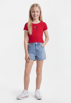 Κορίτσι με μακριά ξανθά μαλλιά, που φοράει κόκκινη ριγωτή μπλούζα με κοντά μανίκια, τζιν σορτς και λευκά ψηλά αθλητικά παπούτσια, στέκεται με το ένα χέρι στην τσέπη.