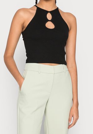 Femme portant un crop top noir sans manches avec deux découpes à l'avant, associé à un pantalon taille haute de couleur claire, main dans la poche.