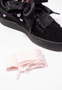 Svart mockaskor med breda svarta satinfjättrar och en strukturerad svart sula, kompletterad med ett långt rosa satinband för alternativ snörning.