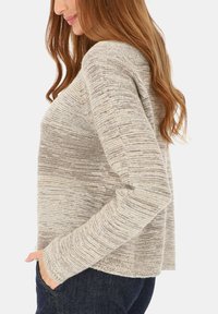 Kobieta z długimi rudymi włosami, ubrana w beżowo-szary sweter dzianinowy i ciemne dżinsy, z lewą ręką w kieszeni, widziana z boku.