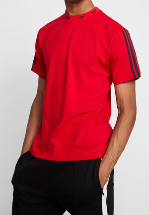 Mand iført en rød t-shirt med korte ærmer og sorte striber på ærmerne samt sorte bukser, hænder i lommerne, foran en ensfarvet baggrund.