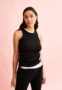 Zwarte geribde tanktop met witte accenten langs de halslijn, gelaagd over een wit shirt. Gecombineerd met zwarte broek, tegen een roze achtergrond.