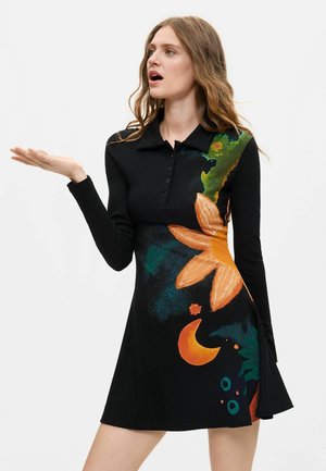 Frau mit langen Haaren, die ein schwarzes Kleid mit langen Ärmeln trägt, verziert mit einer großen orangefarbenen Blume sowie abstrakten grünen und orangefarbenen Mustern.