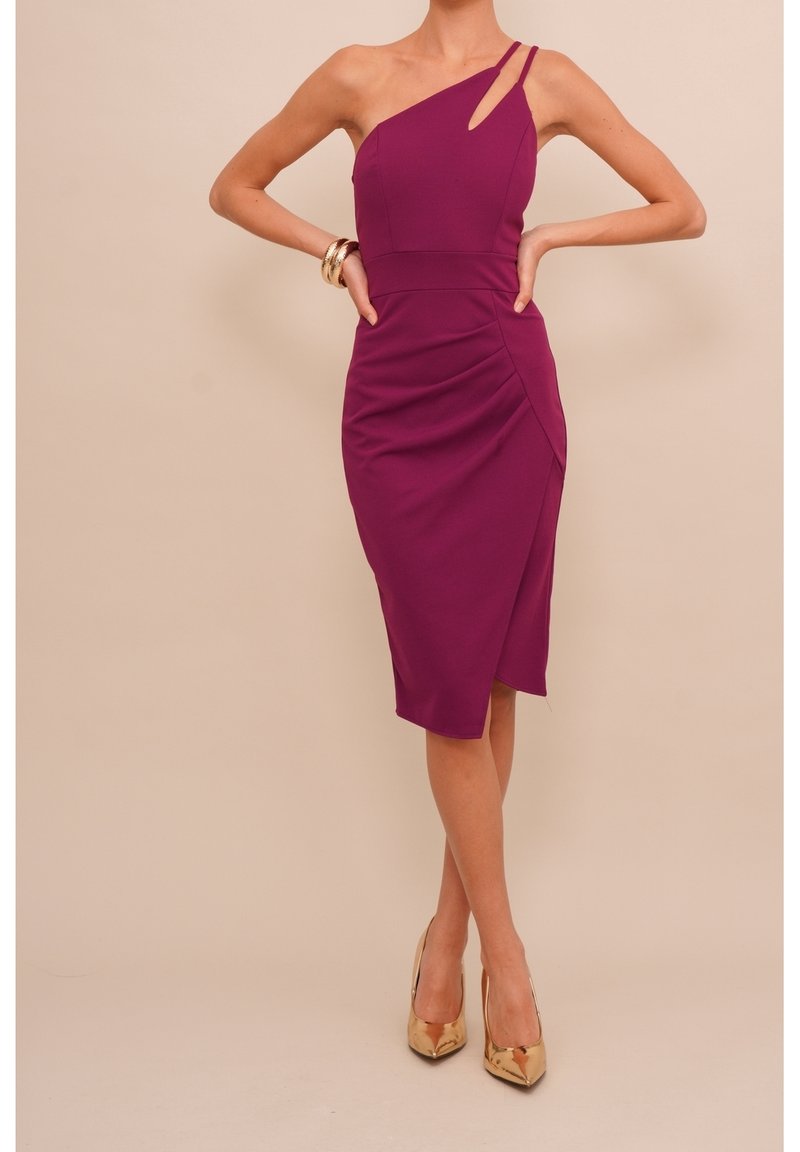 Femme portant une robe violette sans manches aux genoux avec des talons hauts dorés, debout les mains sur les hanches devant un fond uni.