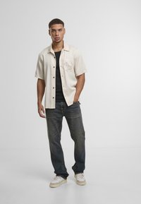 Camisa de manga corta color crema con un bolsillo en el pecho, combinada con jeans oscuros y zapatillas color crema. La camisa presenta un diseño de logotipo sutil.