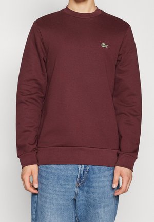 Homme portant un sweat-shirt bordeaux uni à manches longues avec un petit logo crocodile vert sur la poitrine gauche et un jean bleu en denim.