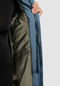 Jakna s podstavom s plavom vanjskom stranom i metalik maslinastozelenim podstavljanjem, sa crnim zatvaračem i elegantnim dizajnom.