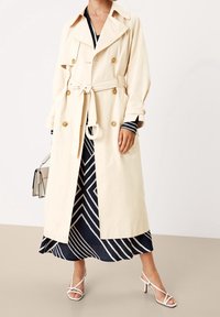 Gabardina beige con botones cruzados, cinturón en la cintura y ajuste holgado. Lleva puesta sobre un vestido azul marino con estampado y zapatos de tacón blancos.